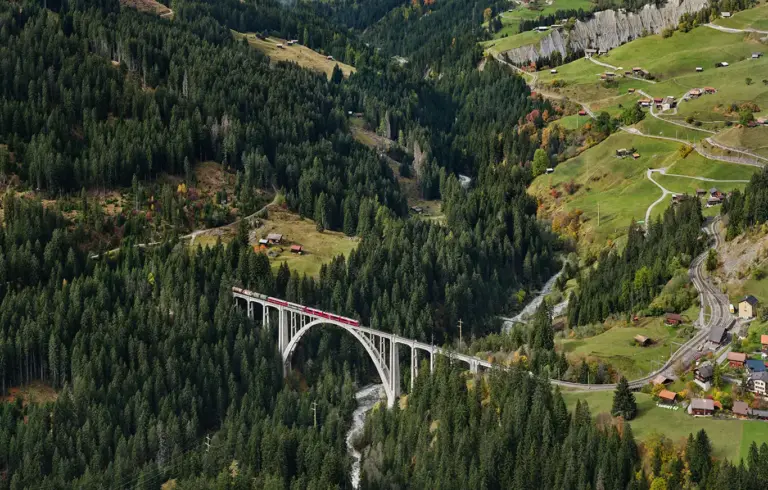 Blick auf den monumentalen, hellen Langwieser Viadukt aus Stahlbeton, der sich in elegantem Bogen über ein tiefes, bewaldetes Tal spannt. Ein roter Zug der Rhätischen Bahn mit angehängten Güterwagen überquert die Brücke