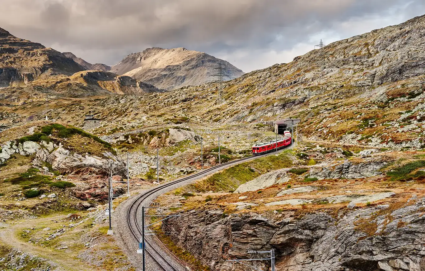 Ein roter Zug der Rhätischen Bahn, angeführt von zwei ABe 4/4 III Triebwagen, schlängelt sich bei letztem Tageslicht durch die alpine Landschaft am Berninapass. Die Schienen führen spektakulär an einem Berghang entlang nahe der Station Alp Grüm. Im Hintergrund erstrecken sich die weiten, rauen Bergketten des UNESCO-Welterbes