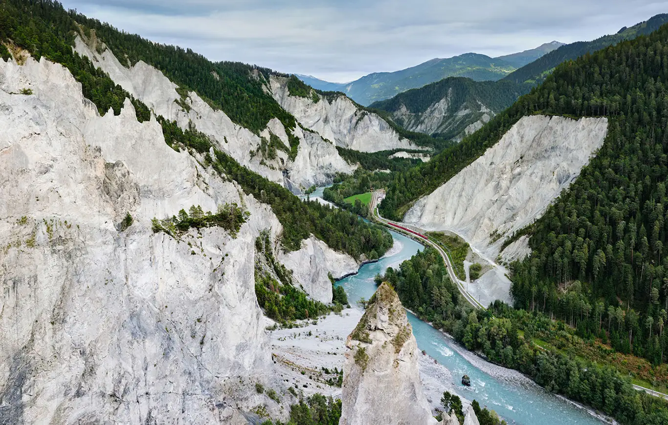 Ein roter Zug der Rhätischen Bahn fährt durch die tief eingeschnittene Rheinschlucht (Ruinaulta). Rechts schlängelt sich der hellblaue, von Kiesbänken gesäumte Vorderrhein durch das Tal, während links die steilen, hellen Kalksteinwände aufragen. Im Hintergrund ist die kleine Bahnstation Versam-Safien inmitten der wilden Naturlandschaft zu erkennen