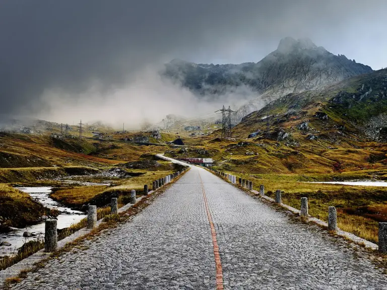 Blick auf die gepflasterte Straße am Gotthardpass bei Nebel. Eine kurze Wolkenlücke gibt die Sicht auf die Straße frei, während die umliegende Berglandschaft mystisch in den Wolken verschwindet