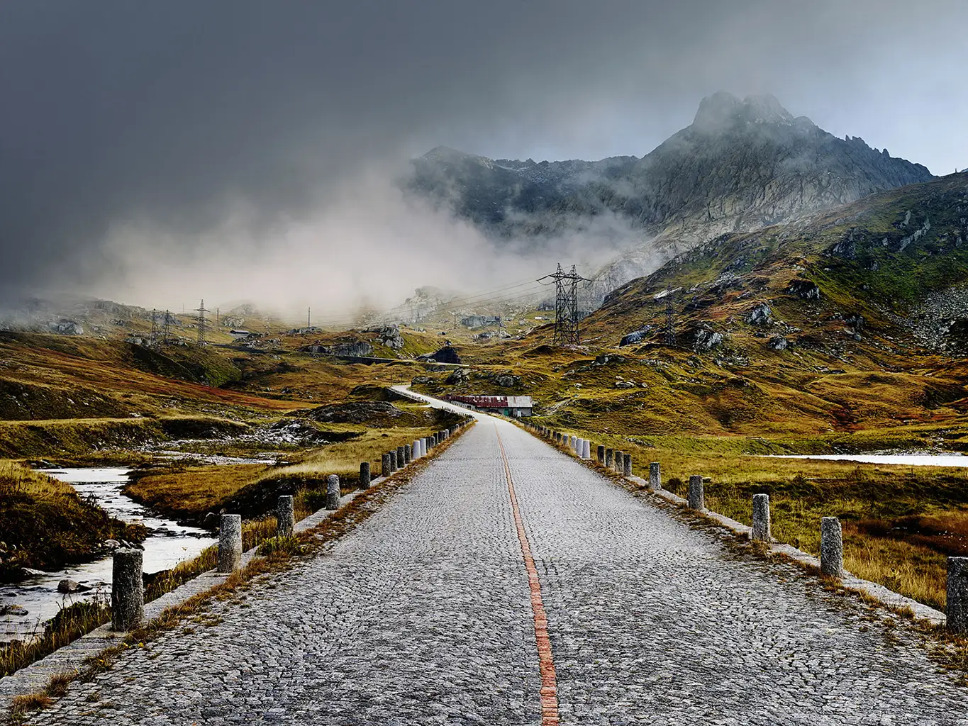 Blick auf die gepflasterte Straße am Gotthardpass bei Nebel. Eine kurze Wolkenlücke gibt die Sicht auf die Straße frei, während die umliegende Berglandschaft mystisch in den Wolken verschwindet