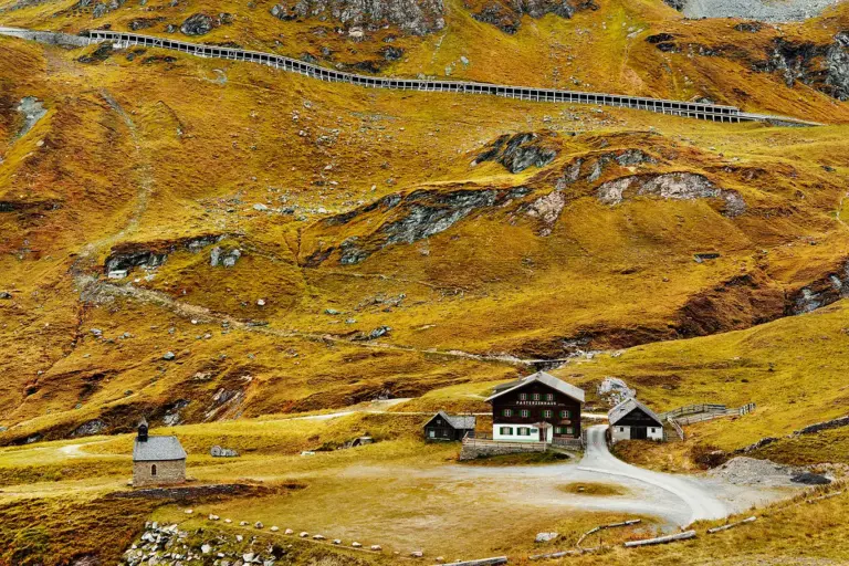 Blick auf das historische Pasterzenhaus und die kleine Herz-Jesu-Kapelle in einer alpinen Hochlandschaft unterhalb der Großglockner-Hochalpenstraße. Im Vordergrund herbstlich gelb-rötlich gefärbtes Gras, im Hintergrund die schroffe Bergkulisse rund um die Kaiser-Franz-Josefs-Höhe