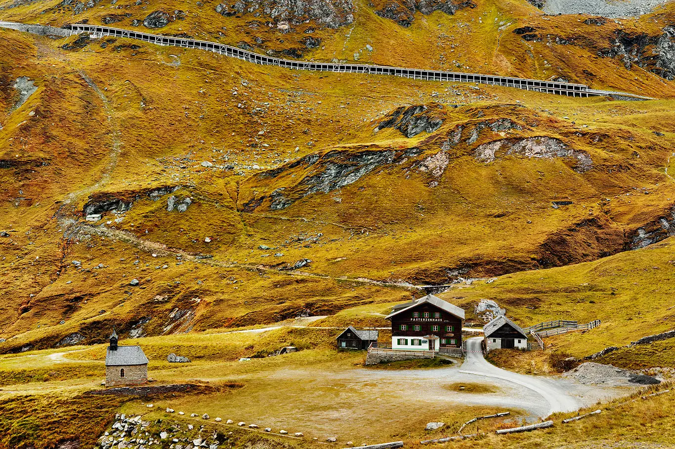 Blick auf das historische Pasterzenhaus und die kleine Herz-Jesu-Kapelle in einer alpinen Hochlandschaft unterhalb der Großglockner-Hochalpenstraße. Im Vordergrund herbstlich gelb-rötlich gefärbtes Gras, im Hintergrund die schroffe Bergkulisse rund um die Kaiser-Franz-Josefs-Höhe