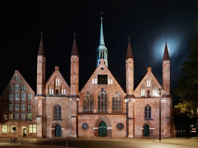 Die beleuchtete Backsteinfassade des historischen Heiligen-Geist-Hospitals in Lübeck bei Nacht. Zu sehen ist die markante dreigiebelige Schaufront mit ihren vier schlanken, spitzen Türmchen, hinter denen der Mond am Nachthimmel schimmert.