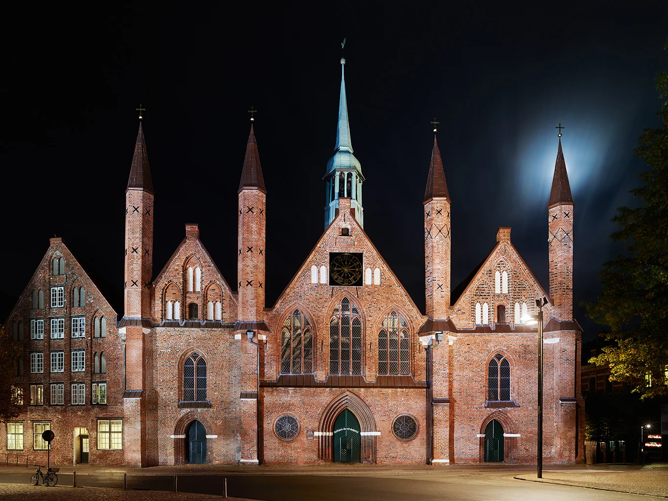 Die beleuchtete Backsteinfassade des historischen Heiligen-Geist-Hospitals in Lübeck bei Nacht. Zu sehen ist die markante dreigiebelige Schaufront mit ihren vier schlanken, spitzen Türmchen, hinter denen der Mond am Nachthimmel schimmert.