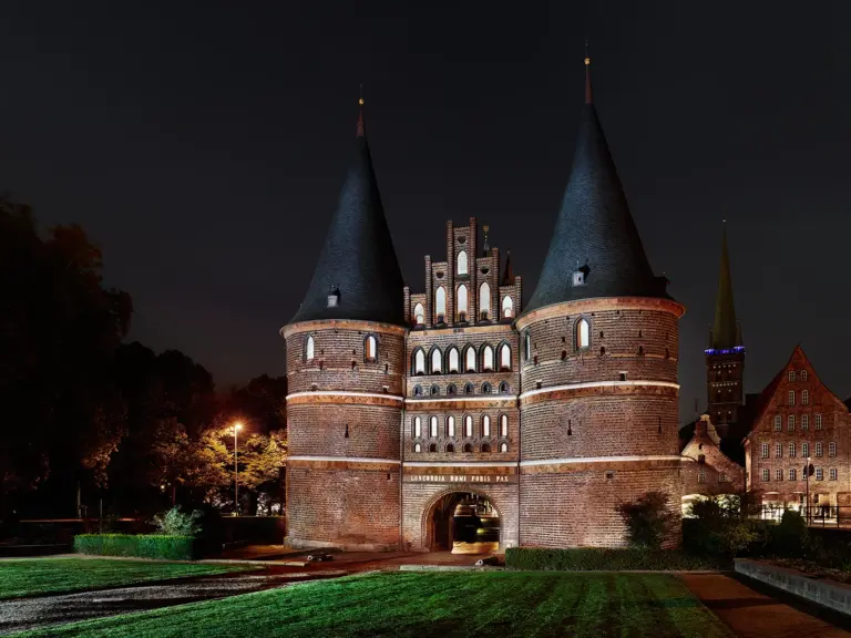 Das historische Lübecker Holstentor bei Nacht, hell erleuchtet vor einem dunklen Hintergrund. Die roten Backsteintürme und das markante Kegeldach spiegeln die typische hanseatische Architektur wider. Deutlich erkennbar ist die historische Schieflage des linken Südturms sowie die lateinische Inschrift über dem zentralen Torbogen