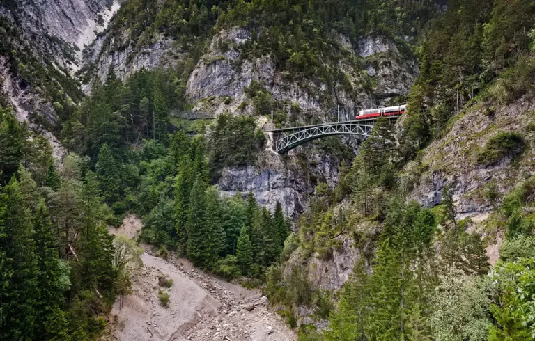 Blick auf die Schlossbachgrabenbrücke, eine eiserne Bogenfachwerkbrücke der Mittenwaldbahn. Die Brücke spannt sich in großer Höhe über eine tiefe, bewaldete und felsige Schlucht. Ein Zug überquert die Brücke vor der Kulisse der schroffen österreichischen Alpen bei klarem Tageslichteichs durch eine der zerklüftetsten Landschaften der Alpen führt