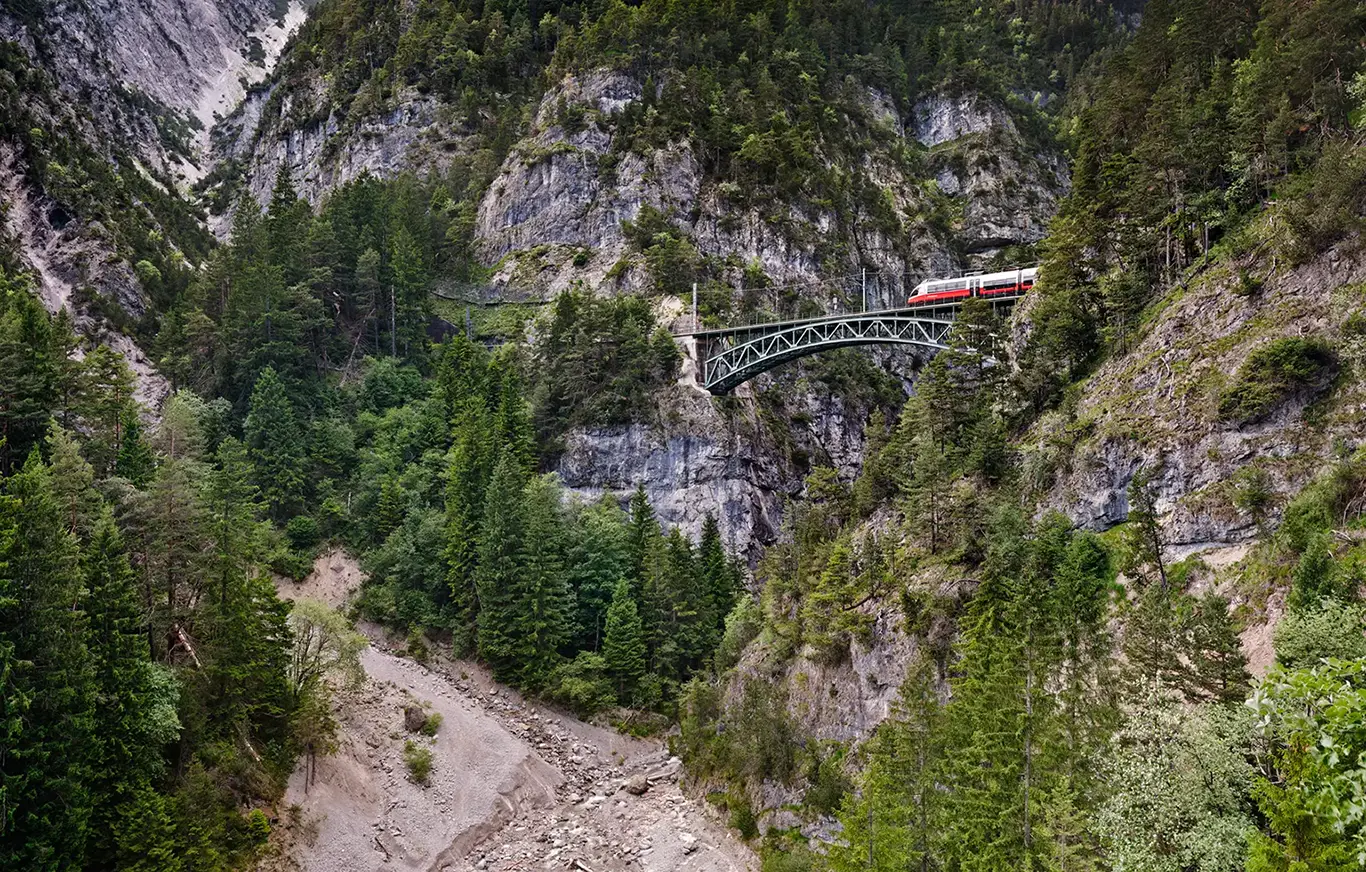 View of the Schlossbach Viaduct, an iron arch truss bridge on the Mittenwaldbahn. The bridge spans at a great height over a deep, forested, and rocky gorge. A train crosses the bridge against the backdrop of the rugged Austrian Alps under clear daylight