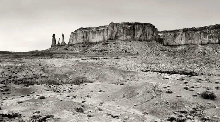 Schwarzweiß-Fotografie der Felsformation „Three Sisters“ im Monument Valley, drei schlanke Sandsteinnadeln vor weitem Horizont.