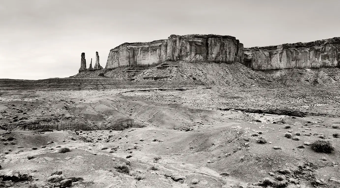 A black-and-white photograph of the ‘Three Sisters’ rock formation in Monument Valley: three slender sandstone spires against a distant horizon.