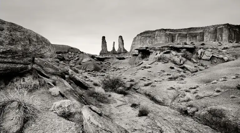 Schwarzweiß-Fotografie der Felsformation „Three Sisters“ im Monument Valley, drei Sandsteinnadeln vor dem Horizont mit prägnantem Vordergrund aus Steinen und Geröll