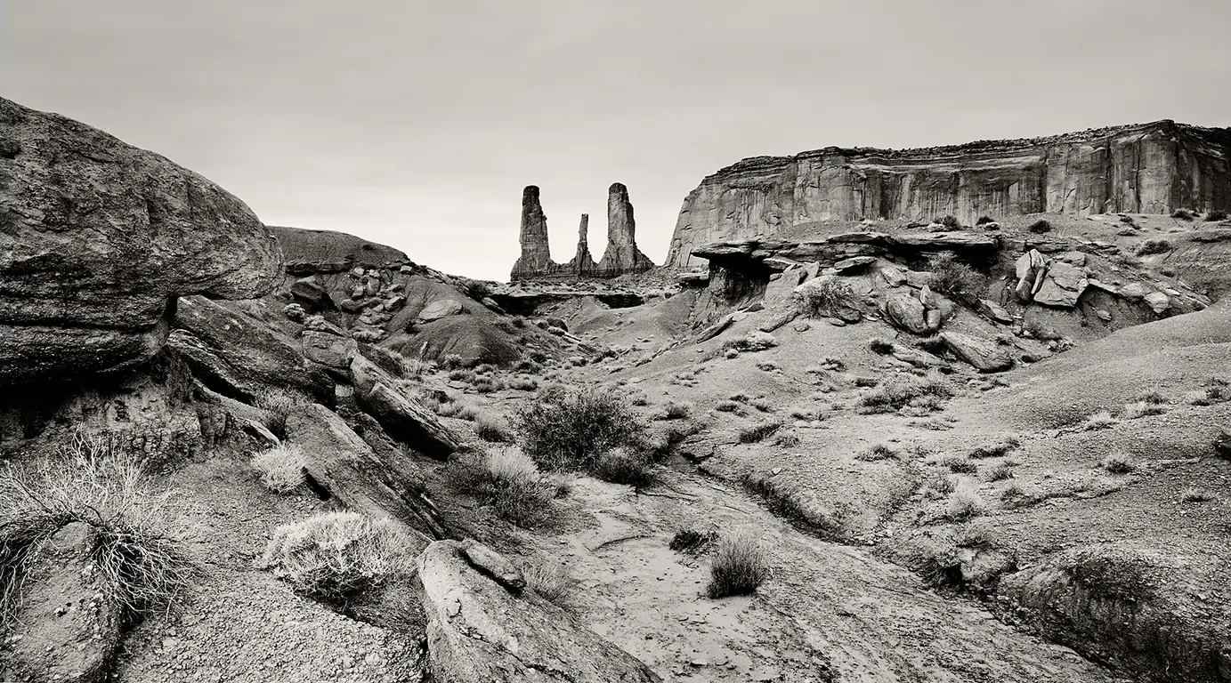 Schwarzweiß-Fotografie der Felsformation „Three Sisters“ im Monument Valley, drei Sandsteinnadeln vor dem Horizont mit prägnantem Vordergrund aus Steinen und Geröll