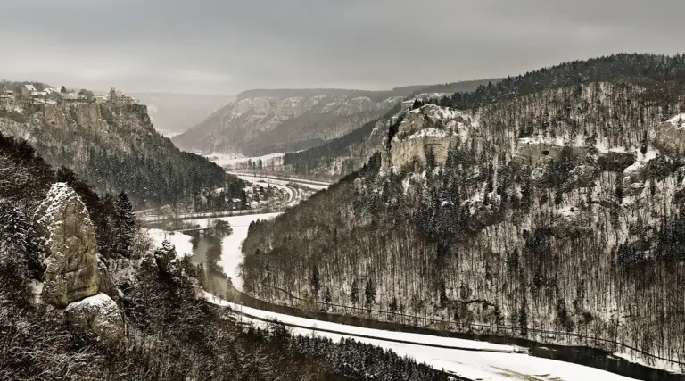 Blick vom Eichfelsen in das tief verschneite Donautal bei Beuron im Spätwinter. Die neblige, gedämpfte Winterstimmung zeigt die kahlen Baumstrukturen, den Flusslauf der Donau und das auf einem Kalkfelsen thronende Schloss Werenwag im matten Licht