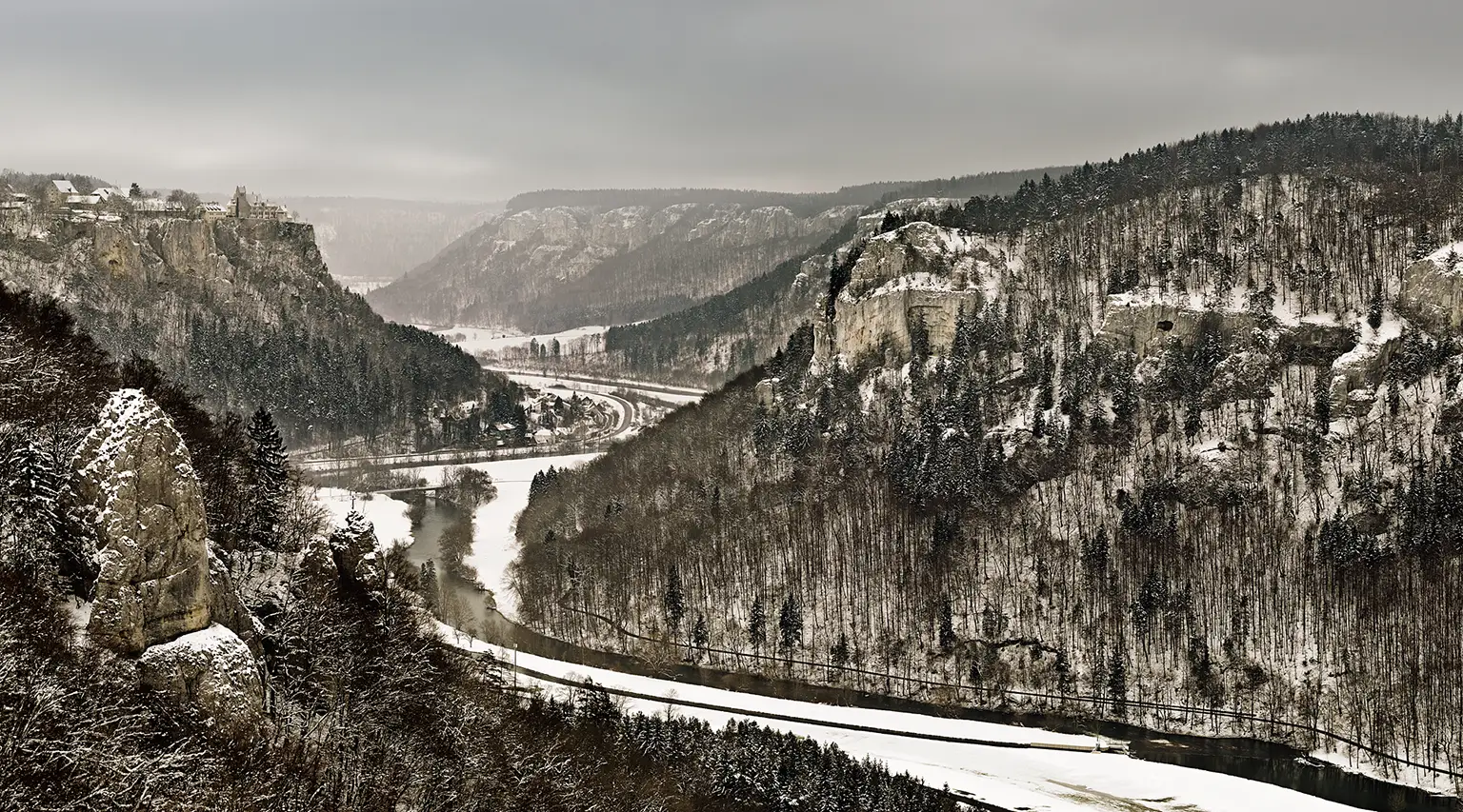 Blick vom Eichfelsen in das tief verschneite Donautal bei Beuron im Spätwinter. Die neblige, gedämpfte Winterstimmung zeigt die kahlen Baumstrukturen, den Flusslauf der Donau und das auf einem Kalkfelsen thronende Schloss Werenwag im matten Licht