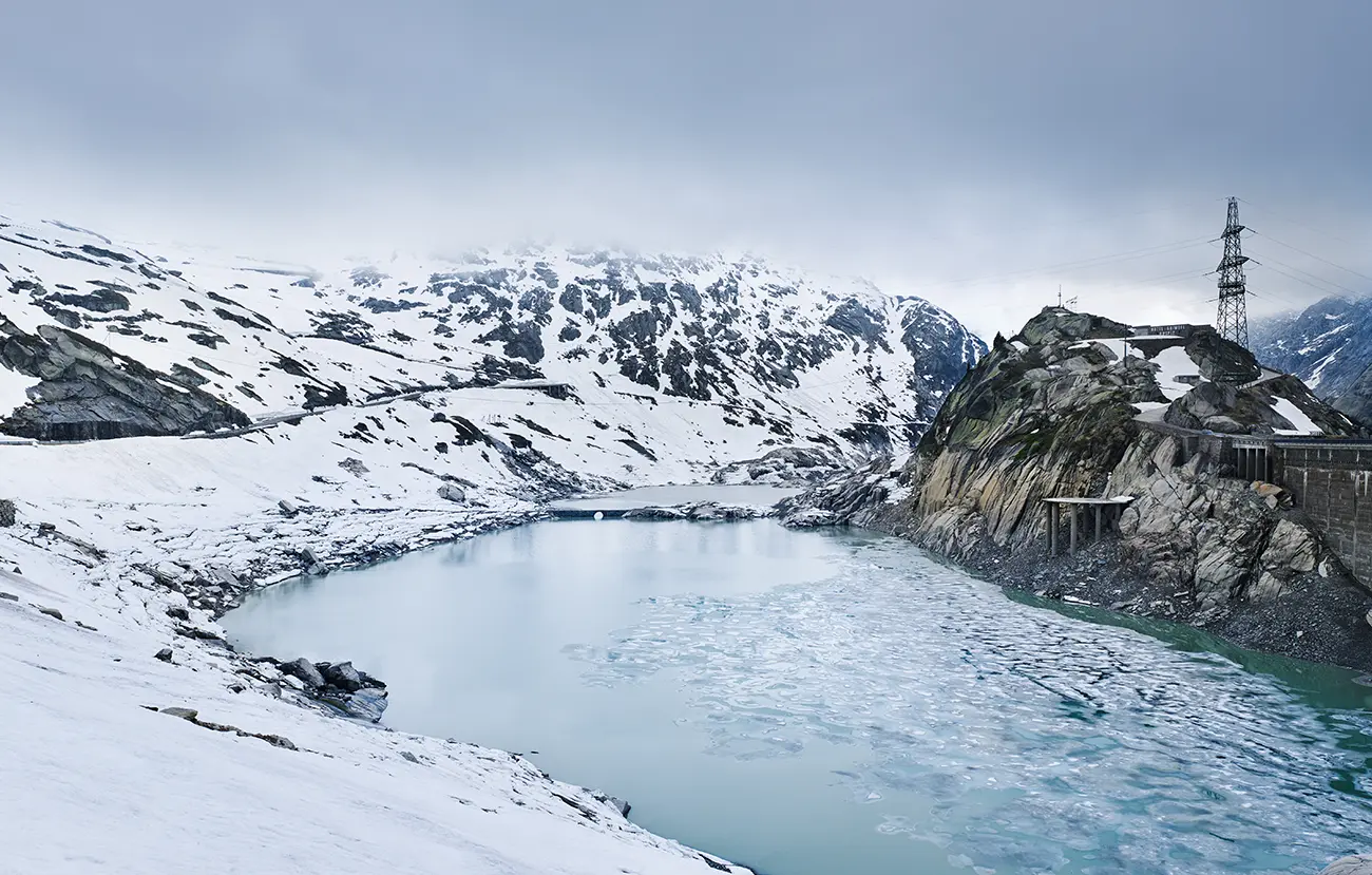 Noch liegt überall Schnee, der Pass gerade erst nach der Wintersperre geöffnet, Wolken hängen tief über den Bergen. Im Vordergrund der Grimselsee, rechts das Hospiz auf seinem Felsen, links die Straße, die sich in Kehren den Hang hinaufzieht. Im See selbst ist noch ein Stück des alten Wegs zu erkennen, mit einer kleinen Brücke aus dem 19. Jahrhundert