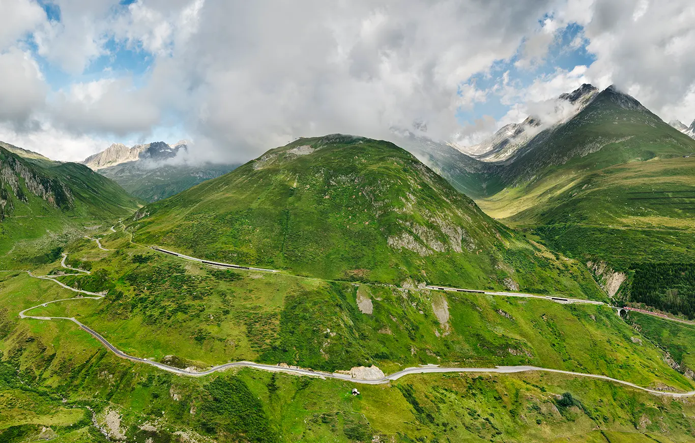 Satt grüne Berglandschaft an der Ostrampe des Oberalppass mit der Bahnstrecke des Glacier-Express und dem Straßenpass