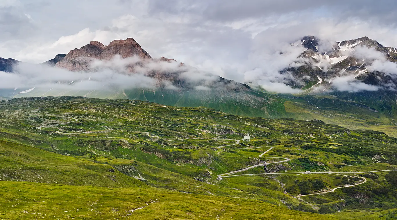 Die Straße zieht sich als feines Band durch die karge Hochgebirgslandschaft, dazwischen der Turm des Lüftungsschacht des Autobahntunnelsaus rohem Sichtbeton